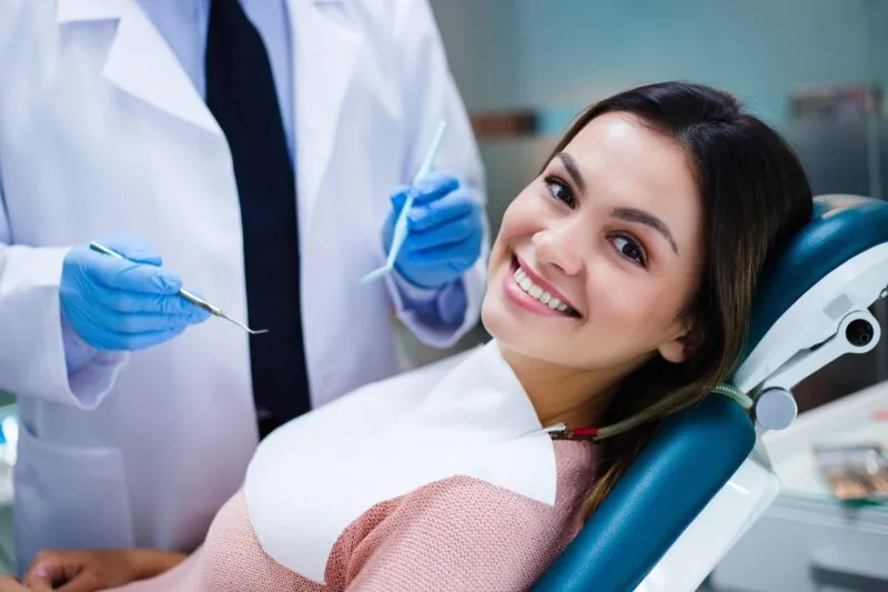 A smiling patient in a dental chair with a dentist holding examination tools nearby.